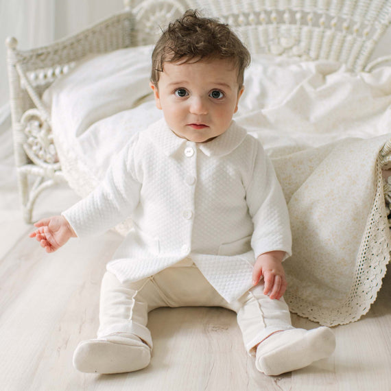 Baby in the Braden Coat Suit sitting on a wooden floor with a white wicker chair and patterned blanket in the background. Perfect choice for high end baby boy suits. 