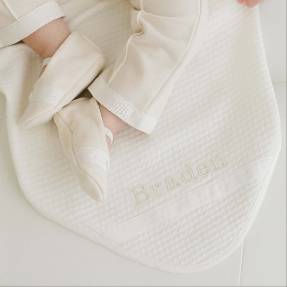 Baby laying on the Braden Personalized Blanket made from ivory textured cotton with an ivory grosgrain ribbon woven on the corner. The name "Braden" is embroidered on the corner of the blanket.