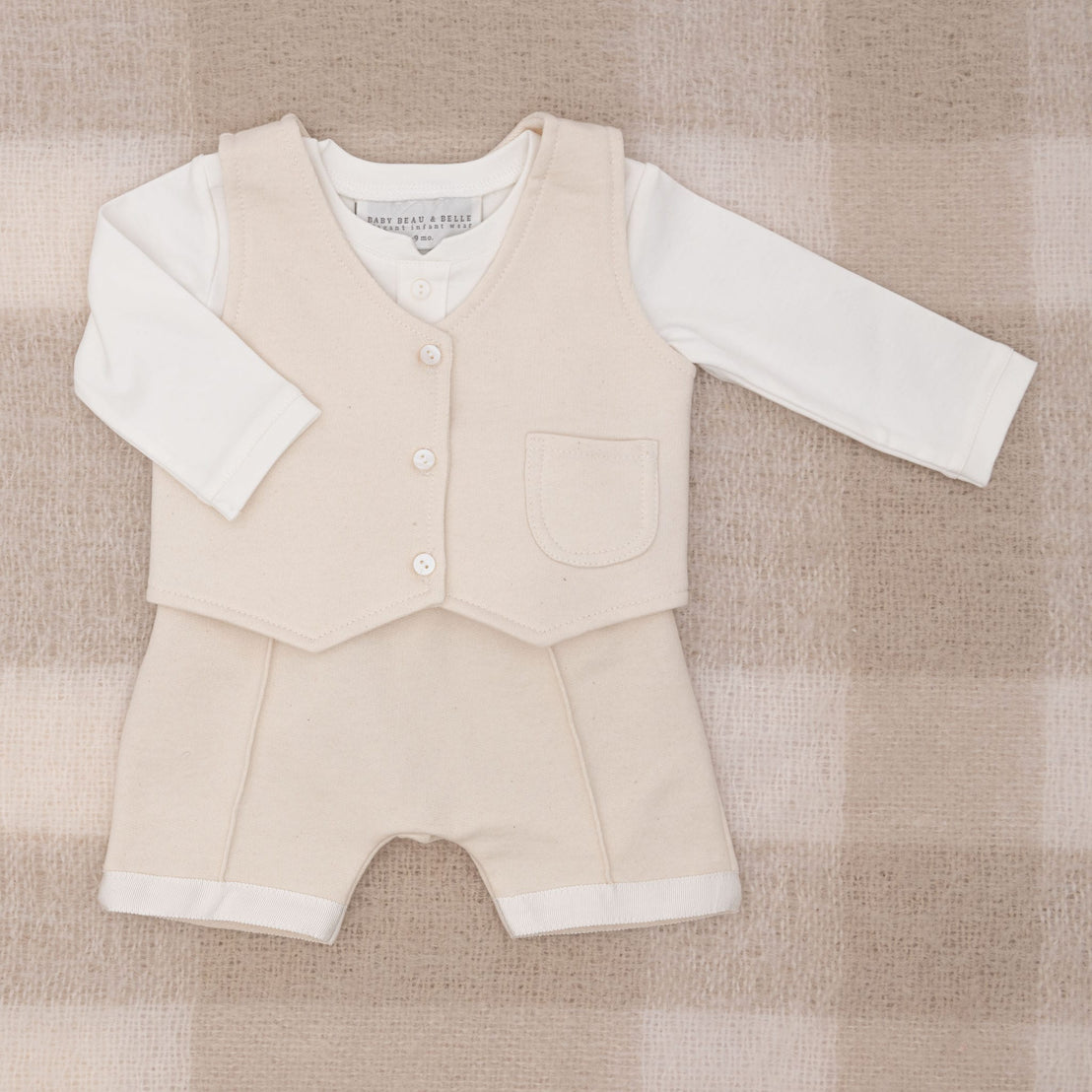 The Braden Vest Suit, a charming baby boy ensemble in beige and white, is displayed on a soft plaid background. This outfit features a cream buttoned vest, a white long-sleeve shirt, and coordinating formal wear shorts made from comfortable French terry fabric.