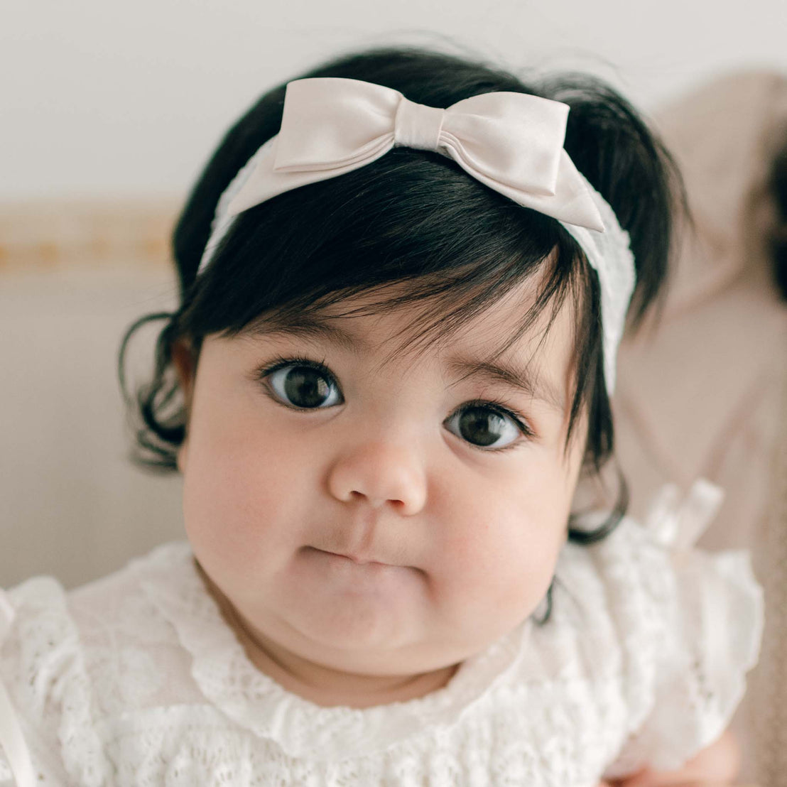 A baby girl wearing the Charlotte Lace Headband made with pink champagne silk and a floral lace. The baby looks directly at the camera with puckered lips.
