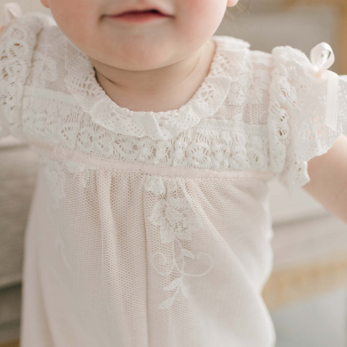 A close-up of a baby girl wearing the Charlotte Romper. The romper features intricate embroidered cotton lace floral patterns and ruffled sleeves. The child’s face is partially visible with a slight smile.
