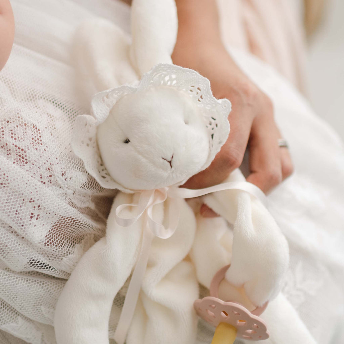A close-up of a soft, white Charlotte Silly Bunny Buddy and Pacifier Holder dressed in a lace bonnet and ribbon. The bunny is being gently held by one hand. The edge of lace fabric and a soft pink pacifier clip are visible in the bunny's hand..