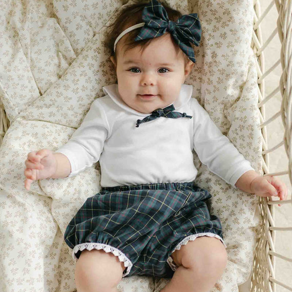 A baby is lying in a woven basket, wearing the Colette Bloomer Set that features a charming white long-sleeve shirt and blue plaid shorts. The little one also sports a matching plaid bow, creating an adorable look perfect for any holiday events. The basket is lined with a patterned cushion, completing this delightful scene.