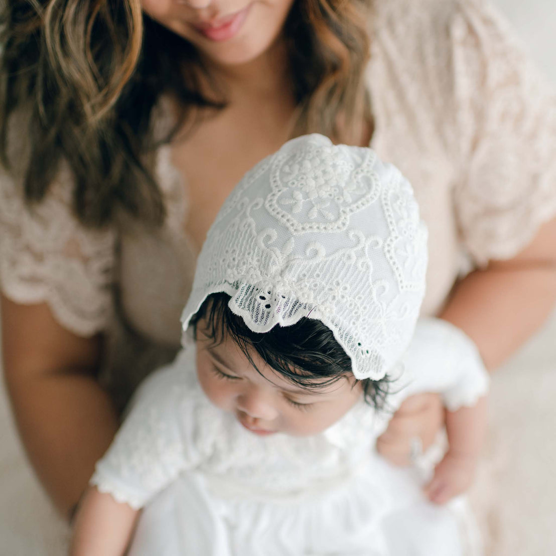 A woman cradles an infant dressed in the Eliza Blessing Gown & Bonnet, crafted from 100% pima cotton, featuring an intricate bonnet design. The baby gazes downward serenely while the woman gently smiles in the background, surrounded by elegant christening accessories with a focus on the child.
