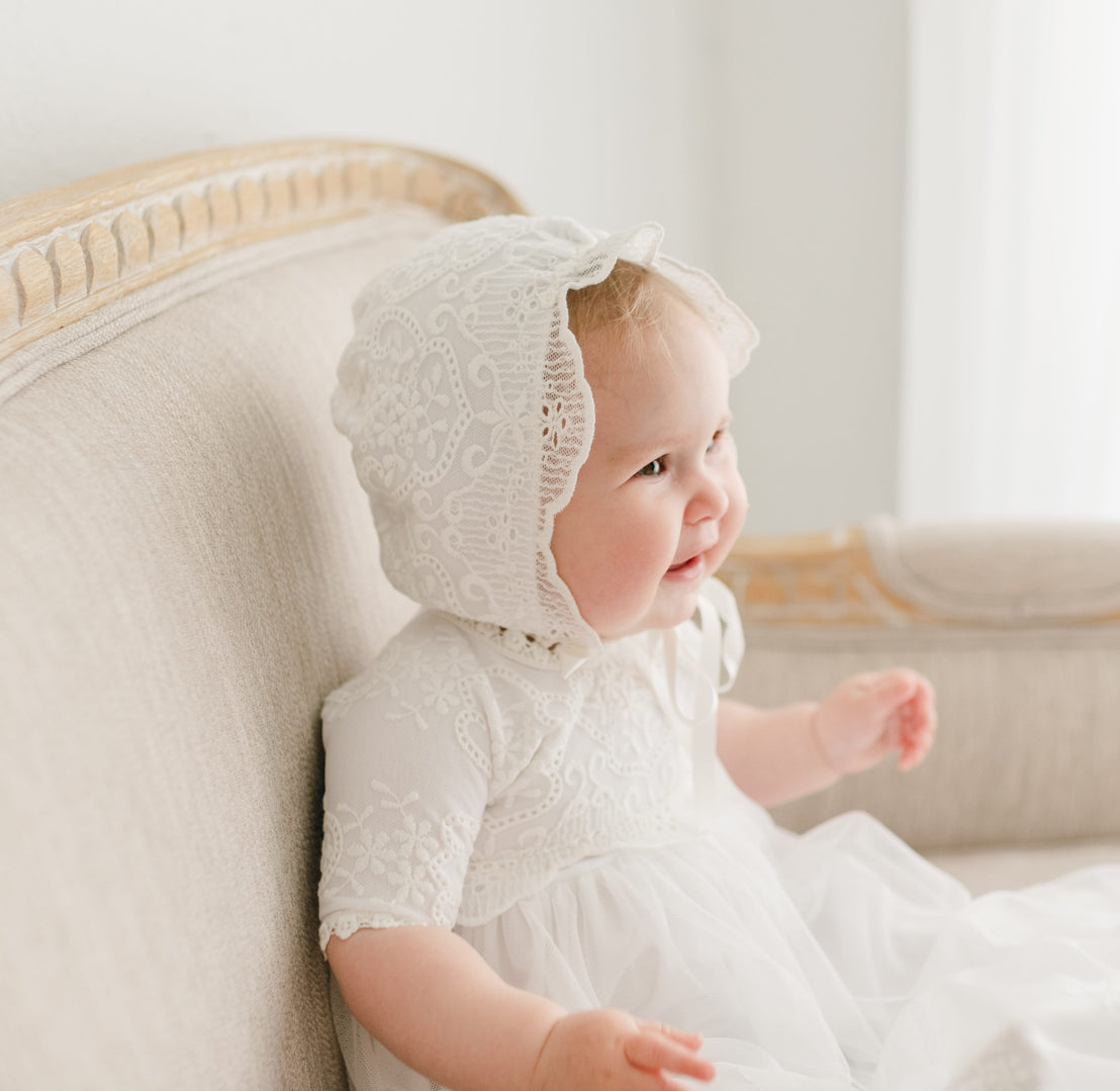 A joyful baby in the Eliza Bonnet and matching gown sits on a beige sofa, smiling and looking to the side with a bright expression.