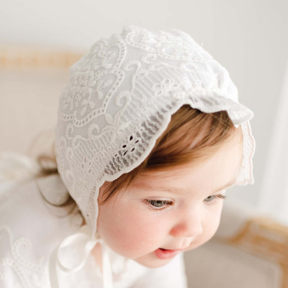 A toddler with light brown hair wearing an Eliza Bonnet and matching dress gazes downward with a gentle expression.