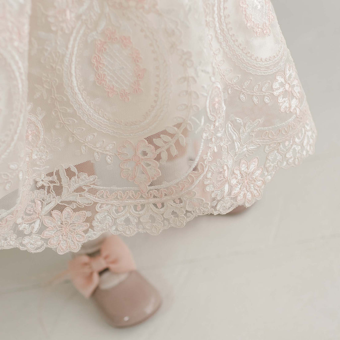 A close-up of the delicate Elizabeth Dress, a handmade baby girl garment adorned with intricate floral embroidered lace and pink accents. Below the baptism dress, a child's foot wearing a shiny beige shoe with a pink bow rests on a light-colored floor.