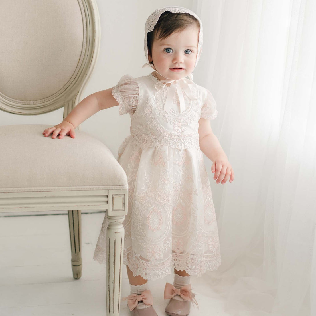 A young child wearing the elegant, handmade Elizabeth Dress, featuring embroidered lace and a bonnet, stands beside a cream-colored chair. The Baptism dress harmonizes with her matching shoes adorned with bows as she looks towards the camera against a soft, light-filled background.