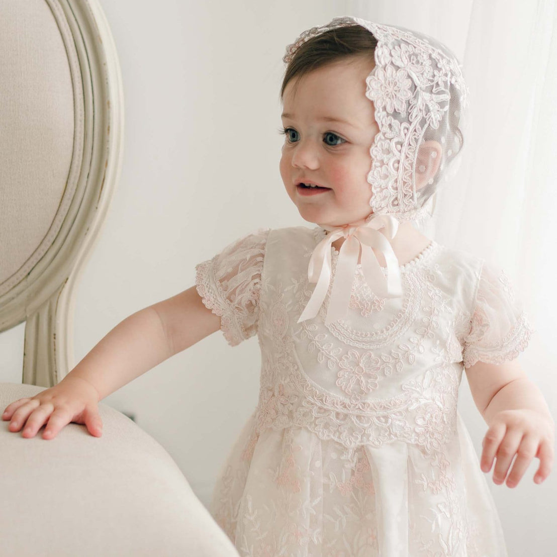 A young child wearing the elegant Elizabeth Dress, a handmade baby girl outfit adorned with embroidered lace and a matching bonnet, stands near a cream-colored chair. The dress is highlighted by its intricate floral embroidery. The child smiles softly as natural light filters through a nearby window.