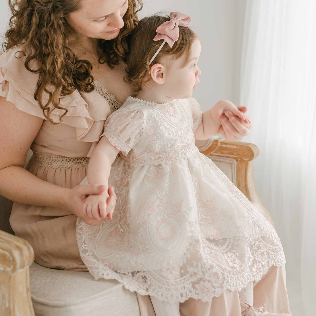 A woman with curly hair sits on a chair, holding a young girl dressed in the Elizabeth Dress, an embroidered lace Baptism gown adorned with a pink bow in her hair. They both gaze toward the window, enveloped in soft natural light. The scene is gentle and serene.