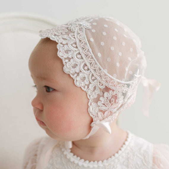 Close-up side profile of a baby in a Elizabeth Lace Bonnet featuring ivory and pink dotted mesh embroidered lace and silk ribbon ties securely fit around her face. Set against a white background. 