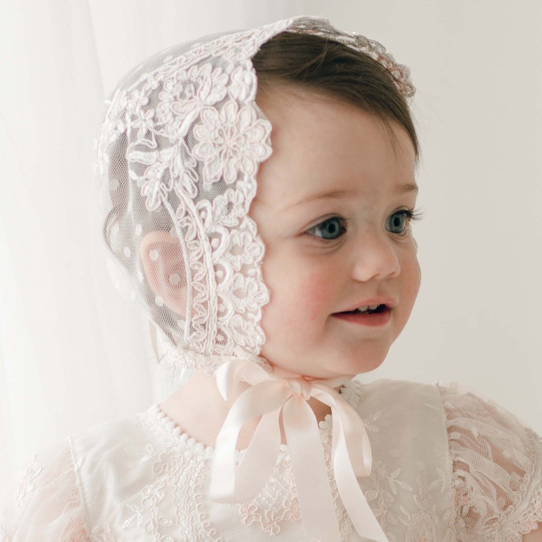 Side profile of a baby girl wearing the Elizabeth Lace Bonnet, featuring delicate floral lace and tied with soft silk ribbons under the chin. The bonnet’s intricate lace detailing frames her face.