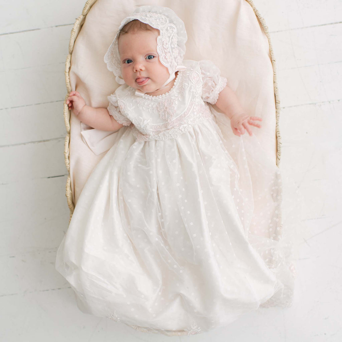 Baby in the Elizabeth Newborn Christening Gown & Bonnet and bonnet lying in a wicker crib on a white wooden floor. Part of the Baby Beau & Belle Baby Girl Christening Dress Collection.