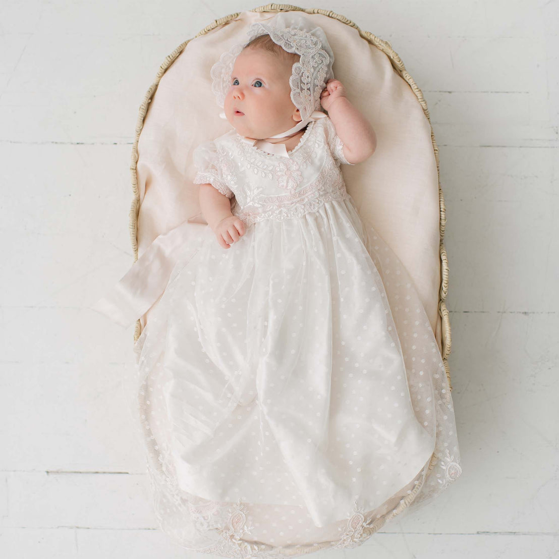 Baby in the Elizabeth Newborn Christening Gown & Bonnet lying on wicker crib placed on a white wooden floor. Part of the Baby Beau & Belle Baby Girl Christening Dress Collection. Perfect for catholic baptism gifts.