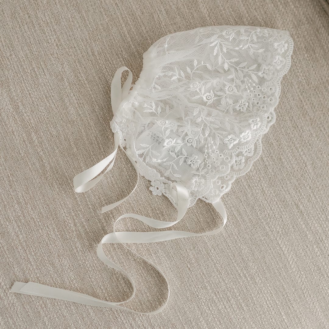A flat lay photo of the Ella lace christening bonnet on a beige fabric chair.