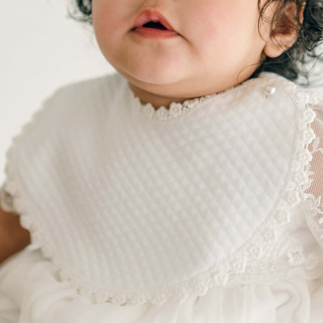 A baby girl with curly hair and expressive eyes wears the Ella Bib and Romper Dress, looking slightly off-camera.