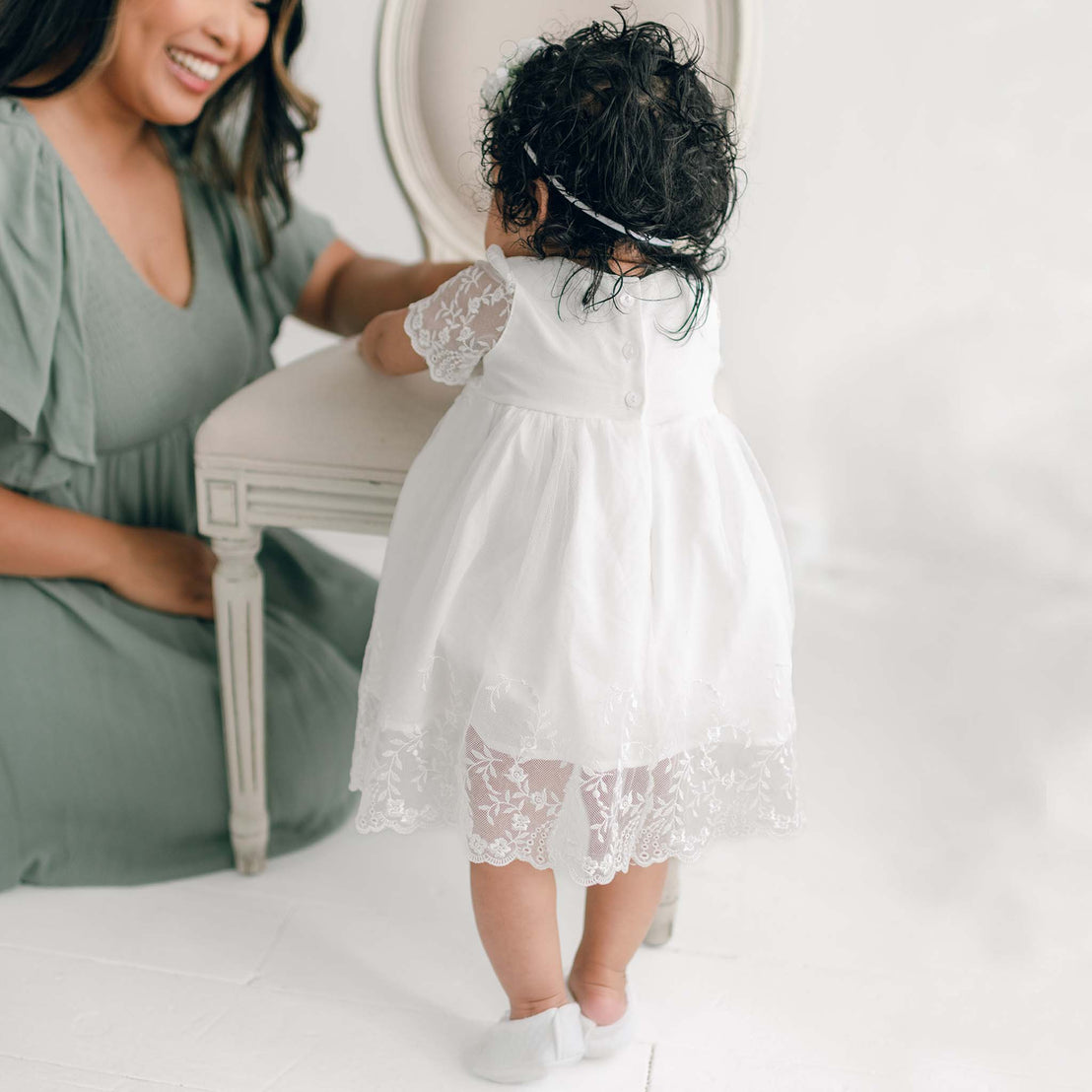 A smiling mother sits beside her daughter in an Ella Romper Dress, who stands next to a white upholstered chair.