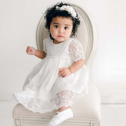 A baby girl with curly hair, wearing the Ella baptism dress flower headband, sits on an light beige chair and looks forward with a curious expression.