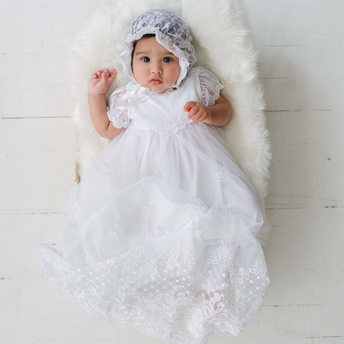 Baby in a white lace dress and bonnet lying on a white rug.