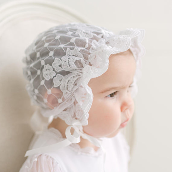 Baby wearing a white lace bonnet against a plain background.