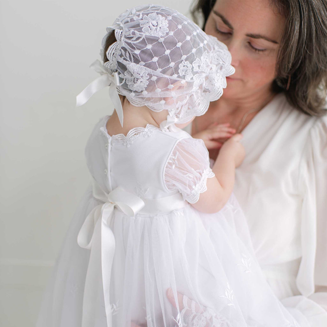 Woman holding a baby dressed in white lace dress against a plain background.