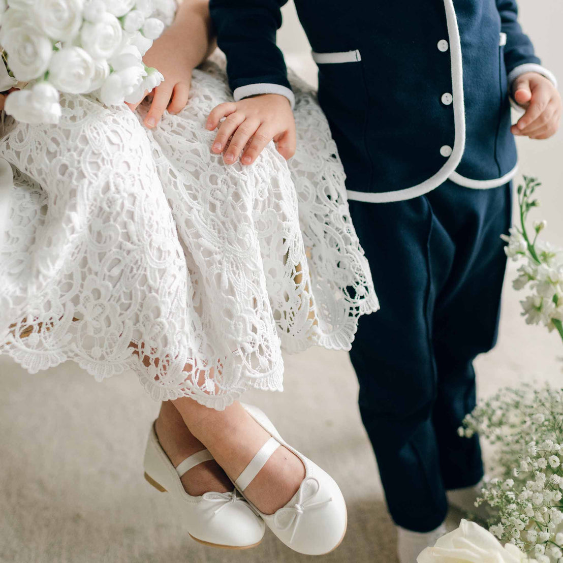 Detail of the Elliott 3-Piece Suit showing the boy’s hand resting on the girl's lace dress near white floral accents.
