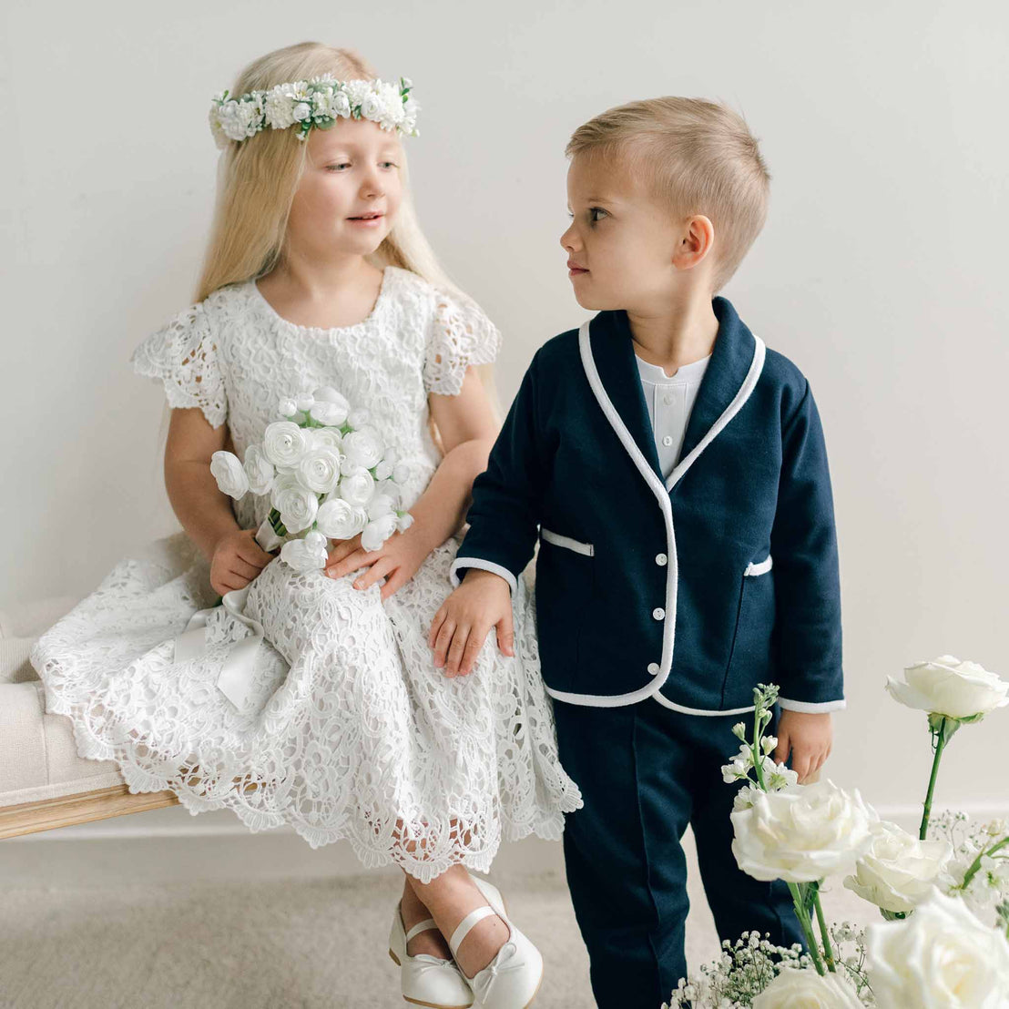 A young boy in the Elliott 3-Piece Suit standing next to a girl in a white lace dress, both positioned near white floral arrangements.
