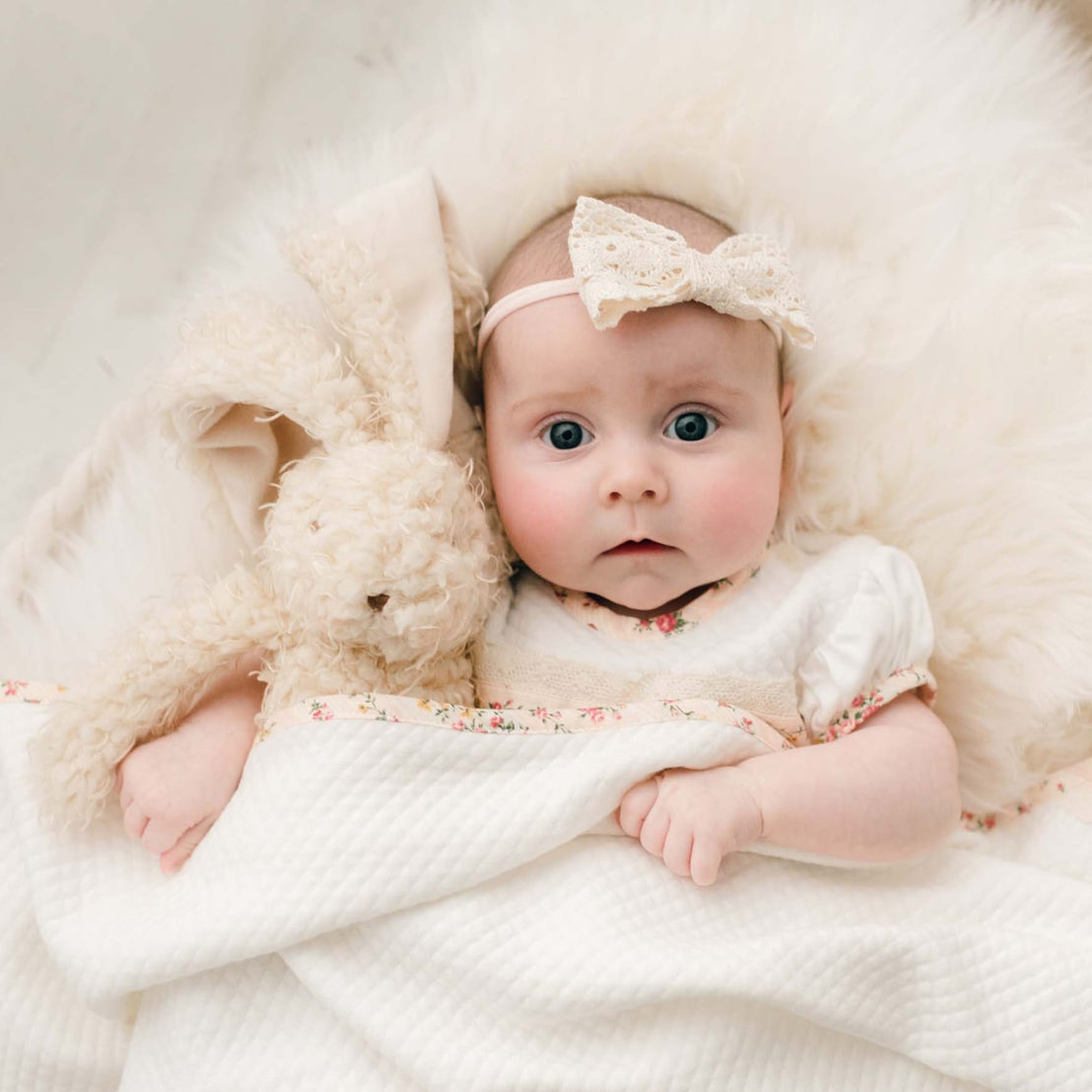 Newborn baby girl underneath a blanket holding the Eloise Bunny, a super soft stuffed animal with a tan scraggly fur body.