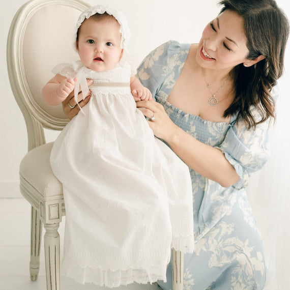 Baby girl with her mother. She is wearing the Emily Gown (with tan sash tie), Bloomers and Bonnet. The Emily Gown features embroidered cotton eyelet lace with a scalloped edge, lace trim and soft lace cap sleeves.