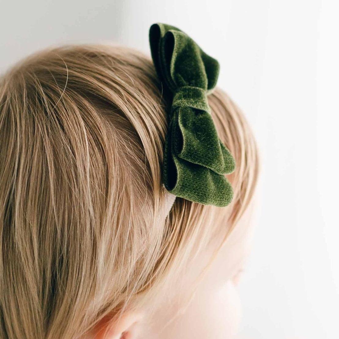 Close-up of a child's head from behind, showcasing short blonde hair neatly styled with an Emily Green Velvet Bow Headband on top for a baptism. The background is a soft white, emphasizing the simplicity and innocence.