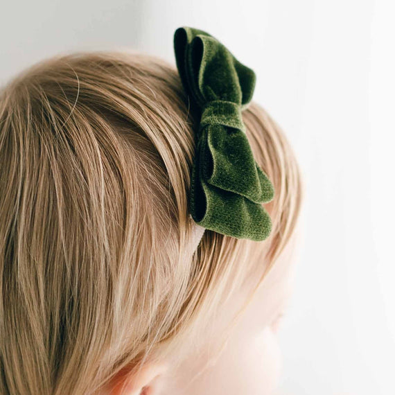 Close-up of a child's head from behind, showcasing short blonde hair neatly styled with an Emily Green Velvet Bow Headband on top for a baptism. The background is a soft white, emphasizing the simplicity and innocence.