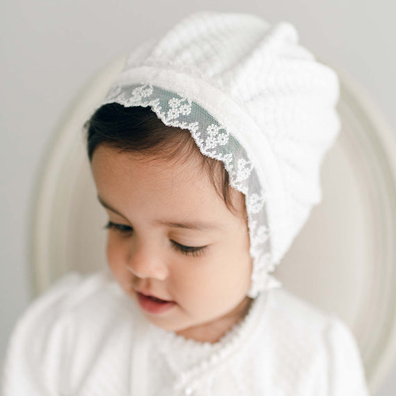 Baby girl gazing down and wearing the Emily Quilted Cotton Bonnet made from a soft quilted cotton in a shade of white and adorned with light ivory floral lace trim.