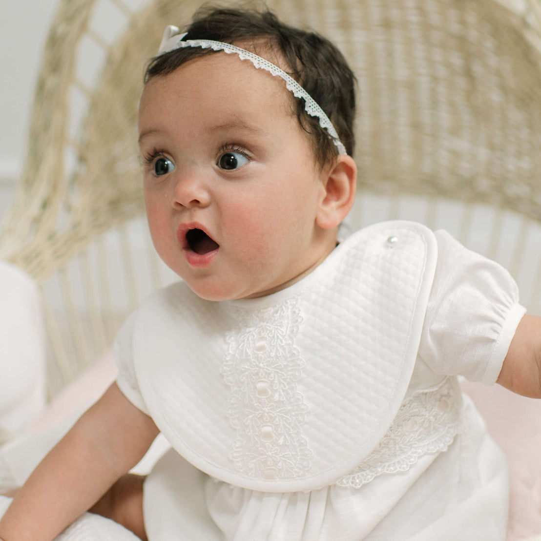 A baby wearing a white long-sleeve dress and a matching white quilted bib with lace trim, sitting in a woven chair. The baby also wears a white lace headband.
