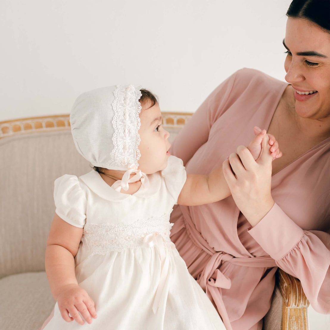 A baby girl wearing the Emma Bonnet and matching dress reaches up to hold her mother’s hand while sitting on a beige upholstered chair. The bonnet features delicate ivory lace trim and silk ribbon ties.