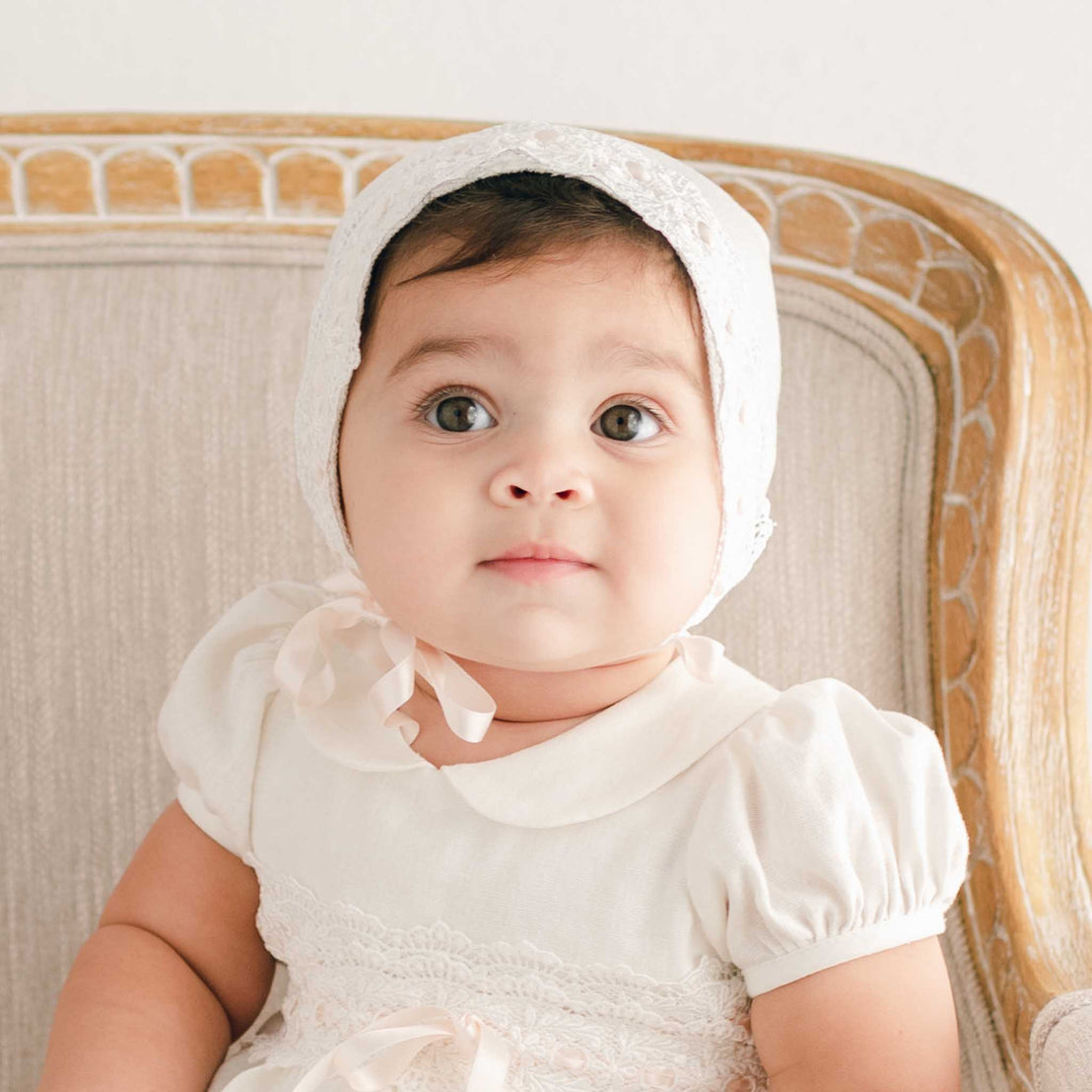 Front view of a baby girl wearing the Emma Bonnet, made from delicate ivory lace and tied with soft silk ribbons, seated on a beige upholstered chair and looking directly at the camera.