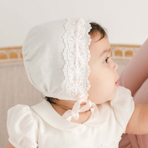 A close-up side profile of a baby girl wearing an Emma Bonnet and matching dress, looking away with a thoughtful expression. The angle showcases the ivory Venice lace along the edge of the bonnet.