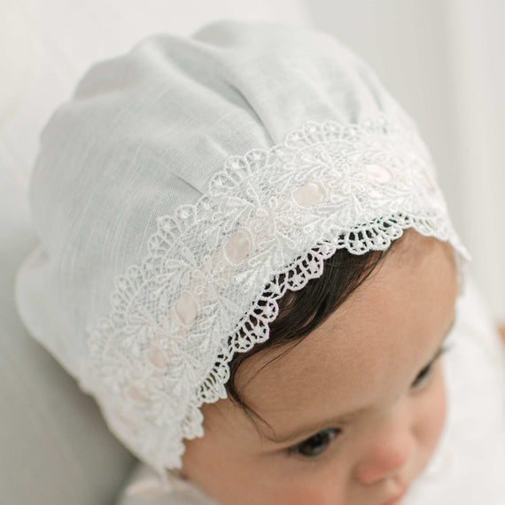 A close-up of a baby wearing a white cotton bonnet with delicate lace trim and small pearl-like accents.