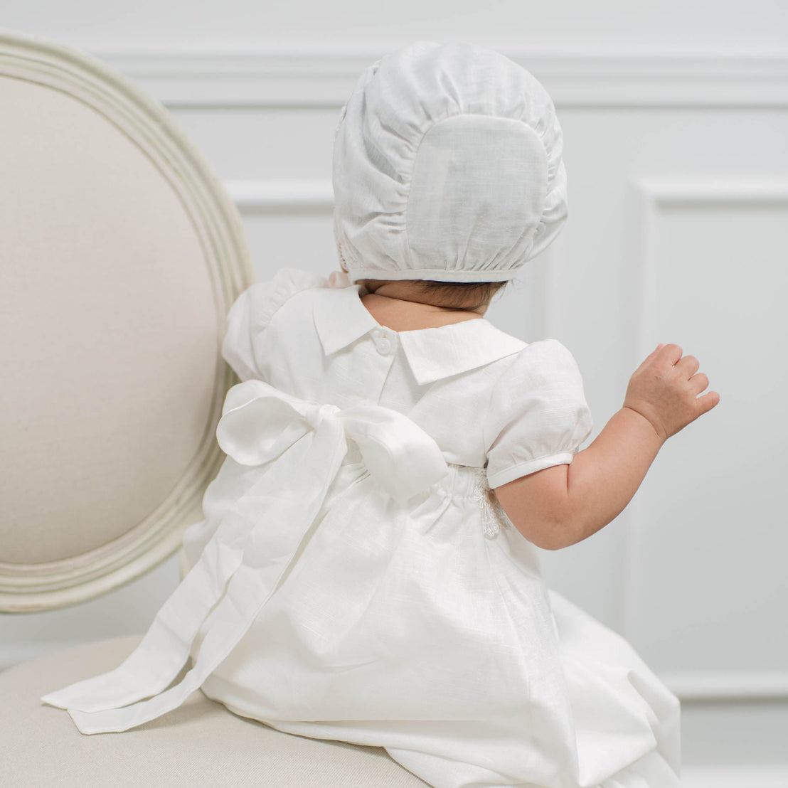 A baby wearing a white short-sleeve dress and bonnet, photographed from behind while sitting on a cream-colored upholstered chair in a bright, neutral room.