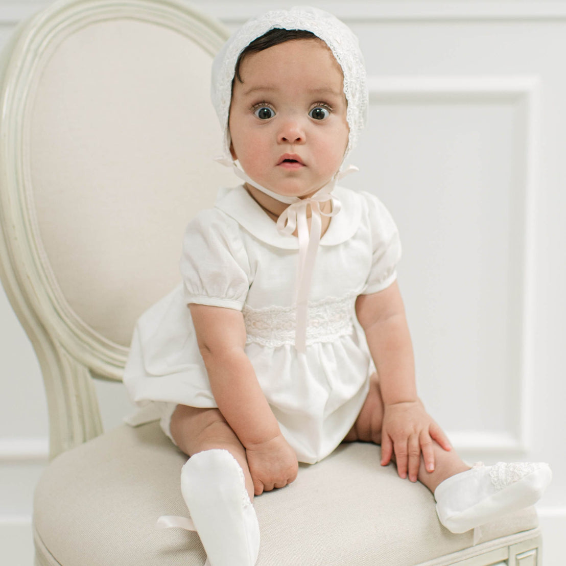 A baby wearing a white short-sleeve dress with lace trim, a matching bonnet, and white booties, sitting on a cream upholstered chair against a white paneled wall.