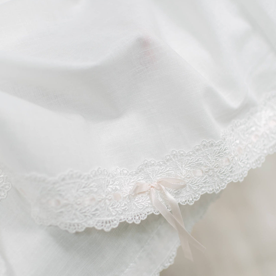 Close-up of a ivory fabric with delicate floral lace trim and a small pale pink ribbon bow at the hem.