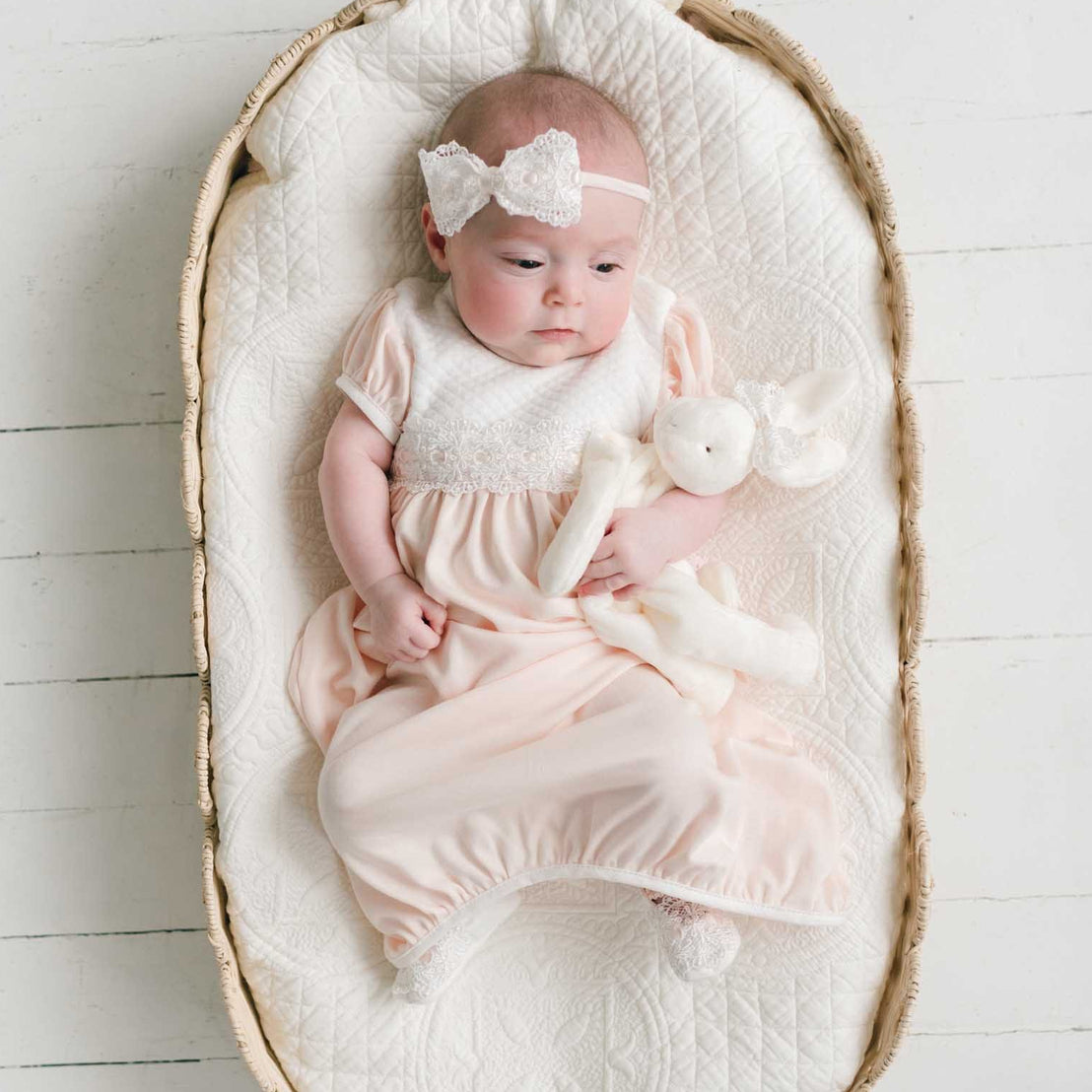 A baby wearing a pale pink footed sleeper and a white bow headband, lying on a quilted cream blanket inside a woven oval bassinet on a white wood floor.