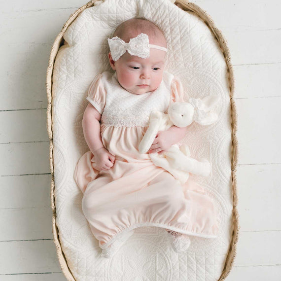 A baby wearing a pale pink footed sleeper and a white bow headband, lying on a quilted cream blanket inside a woven oval bassinet on a white wood floor.