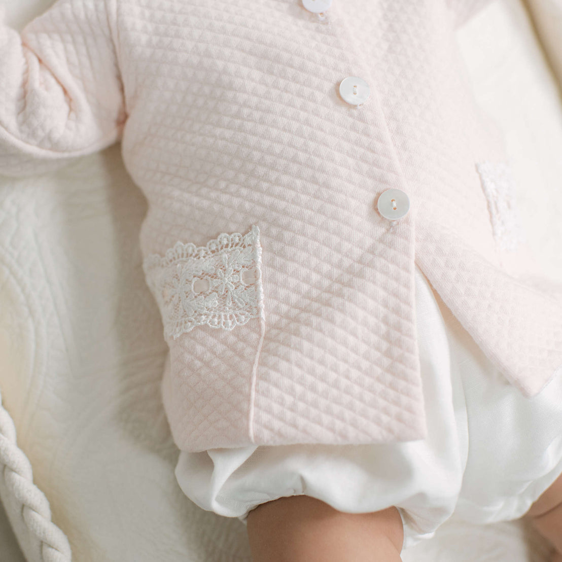 A close-up of a baby wearing a pale pink quilted cardigan with white buttons and a lace pocket detail, paired with white ruffled shorts.