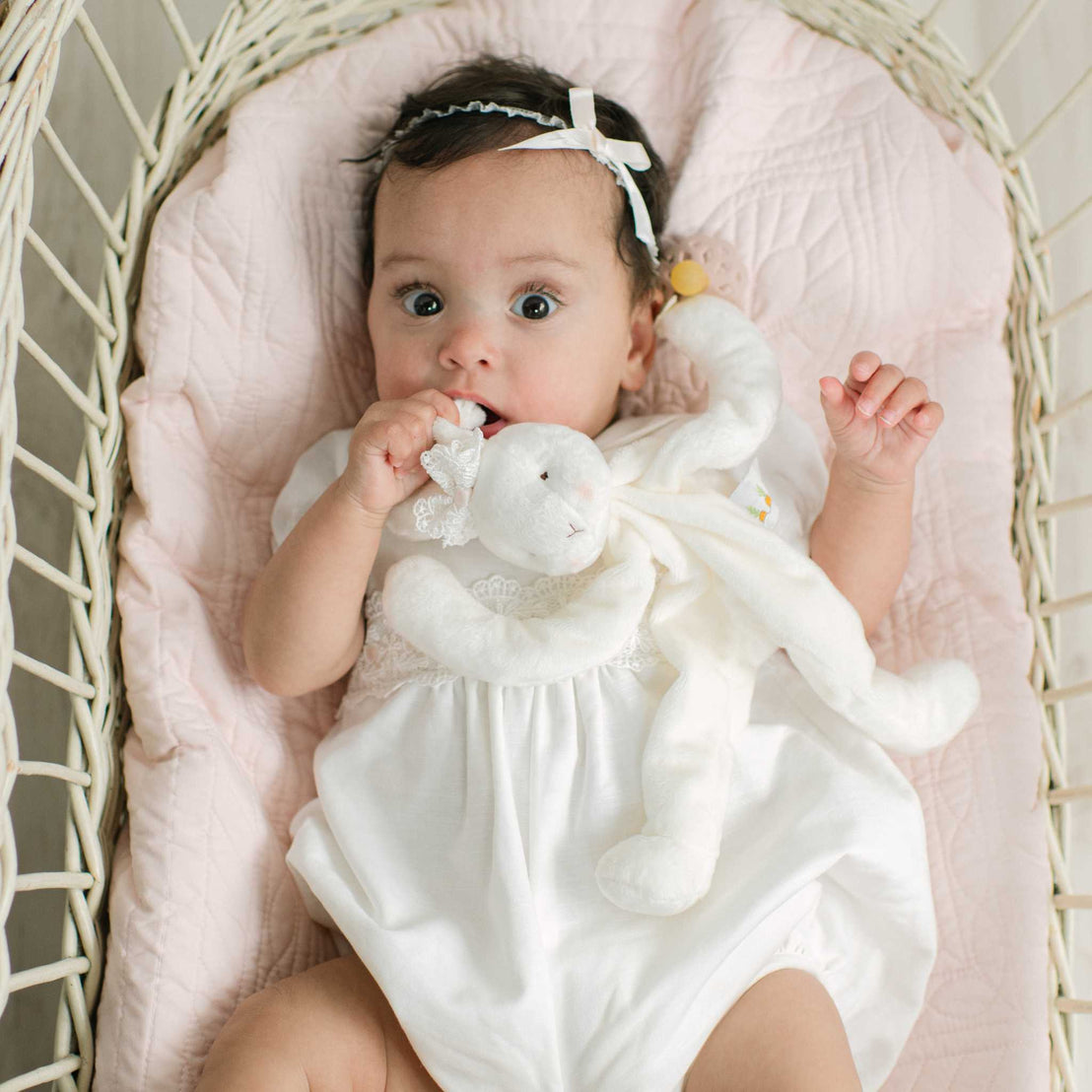 A baby wearing a white dress and matching headband lies in a wicker bassinet on a pink quilted mattress, holding a white plush bunny toy.