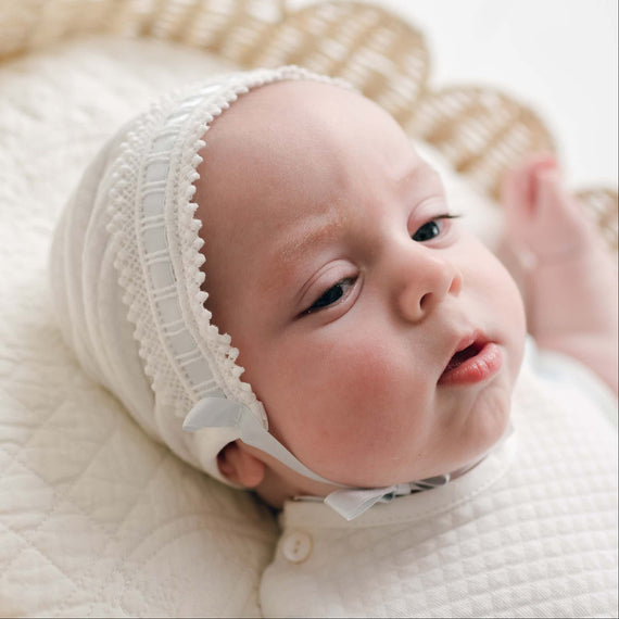 A close-up of a baby wearing an ivory lace-trimmed bonnet and a white quilted swaddle, lying on a cream quilted blanket with a woven basket in the background.