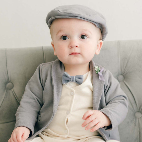 Baby boy sitting on a couch wearing the Ezra 4-Piece Suit with matching Newsboy Cap, heather velvet bow tie and boutonniere
