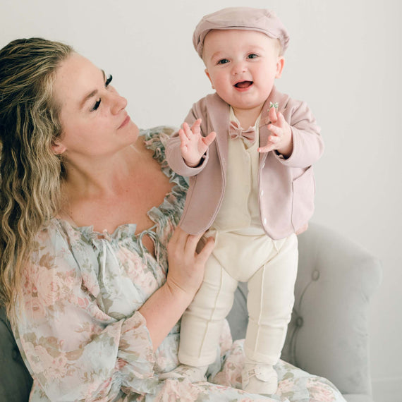 Baby boy standing his mother's lap. He is wearing the Ezra 4-Piece Mauve Suit with matching Newsboy Cap, mauve velvet bow tie and boutonniere