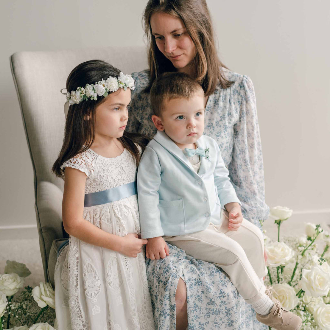 A toddler wearing the Ezra Powder Blue 4-Piece Suit sits on a woman's lap next to a young girl in a lace dress, surrounded by white flowers.
