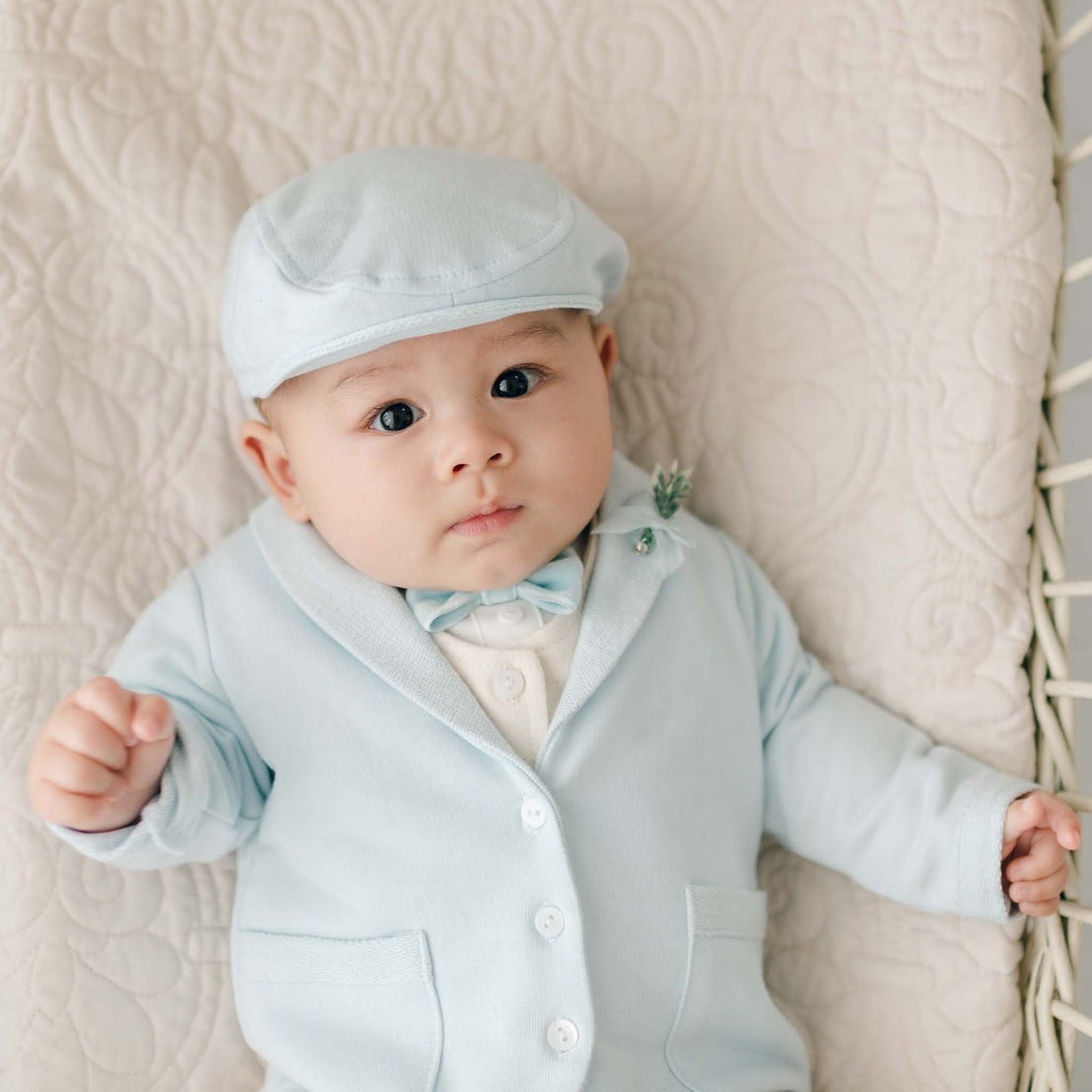 A baby wearing the Ezra Powder Blue 4-Piece Suit and matching cap lies on a quilted blanket, looking at the camera.