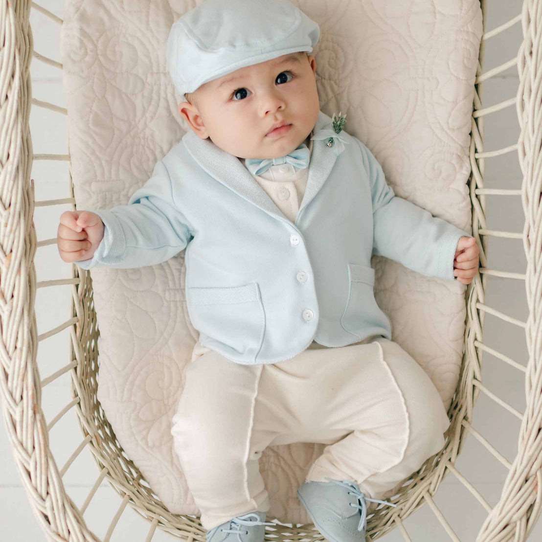 A baby wearing the Ezra Powder Blue 4-Piece Suit and cap reclines in a woven bassinet.